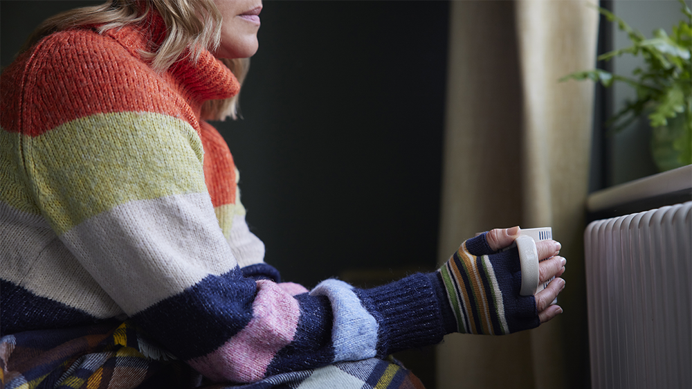 Picture of a person wearing a long-sleeved knit sweater and sitting in front of a radiator. The person is holding a cup and seems to be looking out of a window that is above the radiator.
