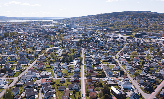An aerial picture of Lillestrøm city. One can see roads, buildings, greenery a hill on the top right side of the picture, and a body of water on the far left side of the picture. 